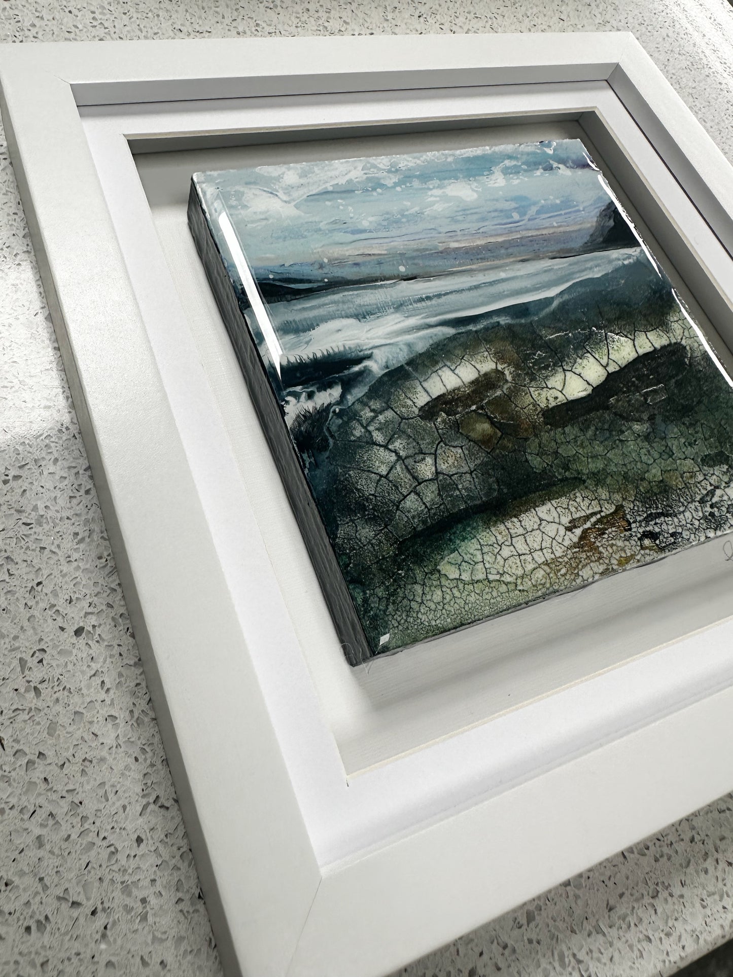 A framed square art print featuring a textured coastal landscape with blue-gray water, rocky shoreline, and cloudy sky, set in a thick white frame on a speckled light gray countertop.