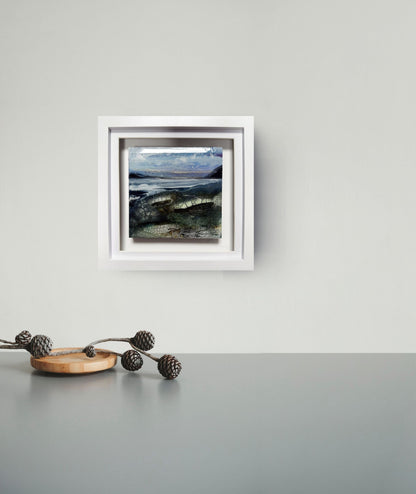 A framed landscape art print hangs on a light gray wall above a dark gray tabletop. The artwork depicts a moody coastal scene with blue-gray water, dark green shoreline, and a cloudy sky, enclosed in a white square frame.